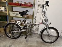 Side view of silver Komda folding bike shows full frame, chain, pedals, wheels, and rear suspension.