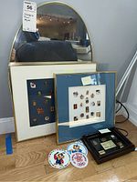 Large oval mirror with gold-tone frame positioned on floor alongside two framed Olympic pin collections and several character cutouts and patch on floor