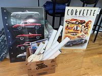 Large Corvette poster leaning against a wall showing various Corvette car profiles in black background beside a box with rolled posters and memorabilia items.