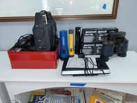 Photo showing various flashlights, a red box with several cords and smaller electronics, and binoculars.