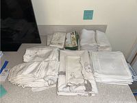Multiple fabric table linens including napkins and covers in white and cream colors with various embroidered and printed designs and neatly stacked on a table.