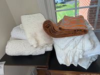 View of multiple white and cream lace table linens with one orange and cream embroidered rolled table runner on top, displayed on a black surface by window light.