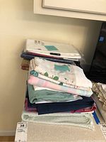 Stack of various folded table cloths and covers including floral, plaid, and solid colors viewed from side angle on a counter.