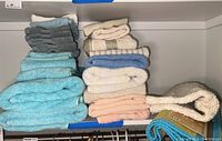 Photo showing multiple folded towels in shades of blue, gray, peach, white, and beige, along with a cream-colored bath mat and a blue trimmed bamboo rug rolled on the side.