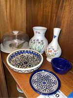 Five ceramic items in wooden shelf: two vases, two ceramic bowls with colorful floral and geometric patterns, small blue bowl, and a cake dome with base visible to the left.