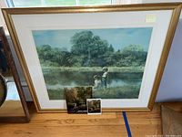 Framed print showing two children by a pond with trees in the background, housed in a gold-colored frame, placed on wooden floor.