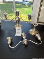 Three sterling silver weighted candle holders displayed on black surface near window: one two-arm candelabra and two single candle holders.