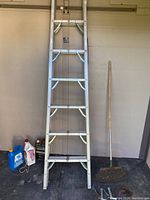 Full view of the 7 ft aluminum folding ladder leaning against wall with plastic containers and a wooden-handled garden rake beside it.