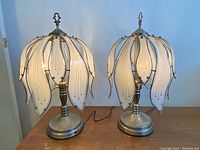 Both antique Deco lamps placed side by side on a wooden surface showing their metal bases and white petal-shaped glass shades.