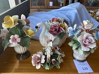 Four porcelain flower figurines with a swan planter, small basket with handle, and two flower vases shown from a front perspective on wooden surface.