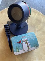 Blue Christmas Star Shower projector with a red power button, red and green stripes, and stake attachment, placed on a table