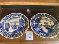 Two blue and white Delft-style plates displayed on a wooden shelf to show entire lot.