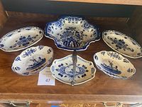 All six Delft blue and white serving dishes arranged on wooden surface showing variety of shapes including oval and scalloped edges, plus one dish with metal handle.
