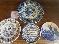 Four blue and white decorative hanging plates arranged on table showcasing traditional Dutch themes including windmill, Dutch woman, floral designs, and Dutch phrase.