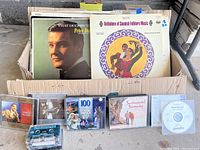 Photo of vinyl records in box plus 6 CDs, 1 cassette tape, and 1 DVD disc laid out in front