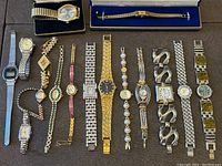 Overview of 15 varied men's and women's wristwatches with metal bracelets laid out on a dark fabric surface.