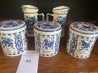 Front view of three Delft blue and white ceramic canisters labeled Tea, Zucker, and Koffie with two matching pitchers behind them on a wooden surface.
