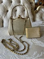 Front view of the gold purse with handle and chain strap beside gold compact on a textured beige cloth surface.
