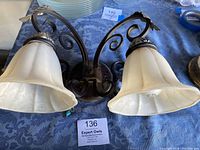Front view of vintage wrought iron wall sconce with two ecru tulip-shaped glass shades on blue patterned tablecloth.