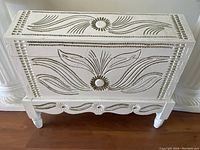 Front view of the white carved wooden hope chest showing floral and leaf carving details and turned legs.