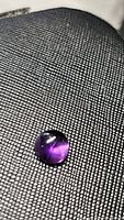 Round deep purple amethyst cabochon gemstone lying on textured black surface, highlighting color and inclusions.