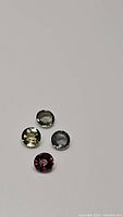 Four loose round cut natural sapphires of mixed colors including green, light yellow-green, bluish gray, and dark reddish purple on a neutral background.