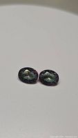 Two oval cut mystic topaz gemstones with purple and rainbow flash colors placed side by side on a plain white background.