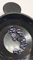 Photo showing sixteen small round polished purple iolite cabochon gemstones in a circular arrangement on a black surface.