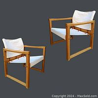 Pair of maple wood frame safari chairs with white canvas sling seats and backs and adjustable leather straps shown against grey backdrop.