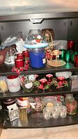 Full cabinet shelf showing multiple Christmas themed candles and candleholders including red and green glass holders, a large blue jar candle with a festive snowman shade, and various decorative pieces including battery-operated candles and berry embellishments.