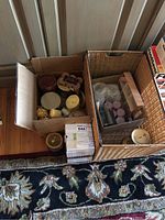 Photo showing multiple candles in a cardboard box and a large woven basket with candle accessories.