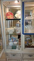 Glass cabinet showing assortment of ceramic bowls, plates, decorative tins and glassware.
