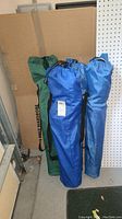 Four folding camping chairs in carry bags standing upright. Three are in blue bags, one in green bag with 'HUGE WARDROBE' text.