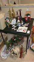 Photo of assorted Christmas figurines, two gold trees, red candle holders, snowman figurines, bird-themed plates, and holiday wreath on a black table.