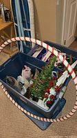 Plastic bin containing small artificial Christmas trees, Christmas figurines, some wrapped in tissue, and a candy cane striped plastic hula hoop leaning inside the bin.