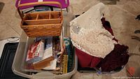 Lot overview showing plastic bin with paper making kit, folded table cloths and doilies, and woven basket on tiled floor.