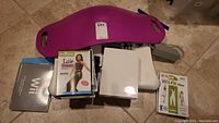 Photo showing purple Simply Fit Board with handles, Nintendo Wii game cases including Latin Grooves, Wii Fit Plus, and instruction manuals spread above it on floor.