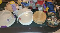 Photo showing white plastic cake carriers with dome lids, woven round trays, plastic cups, mugs, cutlery in holder, an M&M candy dispenser, and additional kitchen and party items on a dark table.
