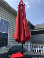 Closed red patio umbrella installed in metal stand outside on a black table with deck and house siding in background.