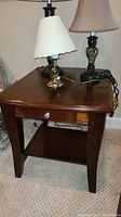 Full view of the wooden side table showing the top surface, drawer, lower shelf, and legs, along with two lamps on top.
