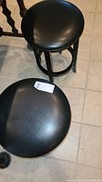 Top-view and side-view photos showing two black round bar stools with vinyl seats and wooden legs, one stool has a crack on the base.