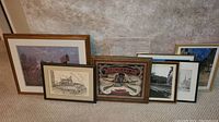 Eight framed pieces of art leaning against wall on carpeted floor showing different sizes, styles, and themes