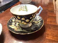 Front angled view of tea cup filled with saucer showing floral pattern on black background with gold edging