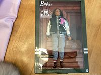 Photo of Barbie x Roots Limited Edition doll in original packaging, showing doll dressed in varsity jacket, grey pants, brown shoes.