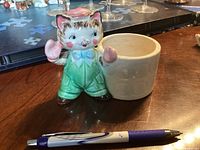 Front view of ceramic cat figurine wearing green overalls, blue bow tie, and pink boxing gloves, next to white pot