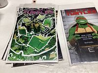 Five Teenage Mutant Ninja Turtles comic books in clear protective sleeves. Left comic shows Leonardo surrounded by green lightning, right comic shows Raphael in police lineup.