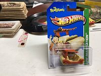 Photo of the 2012 Hot Wheels Flintstones Flintmobile toy car in original packaging showing a blue card with Hot Wheels and The Flintstones branding, featuring Fred Flintstone illustration, and the tan and brown Flintmobile inside clear blister.