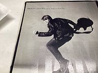 Front cover of Bryan Adams 'Cuts Like a Knife' vinyl record showing black and white image of Bryan Adams in mid-air pose holding a guitar.