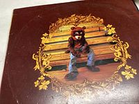 Front cover of Kanye West The College Dropout album showing bear mascot sitting on wooden bleachers with gold ornate frame design on cover.