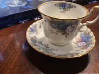 Tea cup and saucer with blue rose floral design, gold rim and handle, placed on wooden surface.